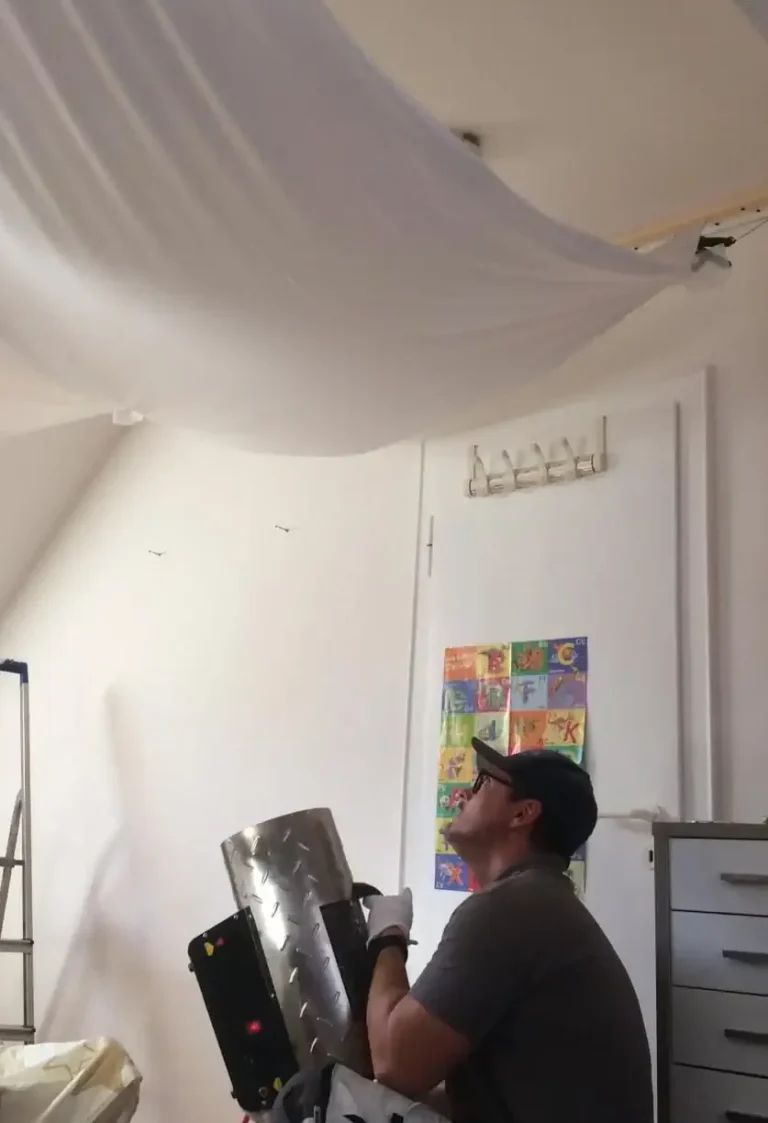 Installation einer Spanndecke im Schlafzimmer. Die Decke ist aufgehangen und wird zum Einspannen erwärmt. Mann hält einen Heizlüfter.